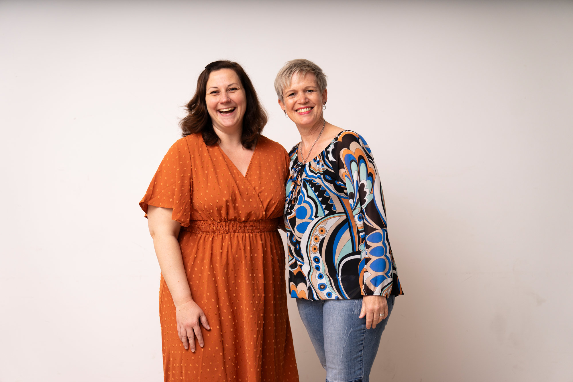Zwei lachende Frauen, eine im orangefarbenen Kleid, die andere in bunter Bluse und Jeans, posieren.